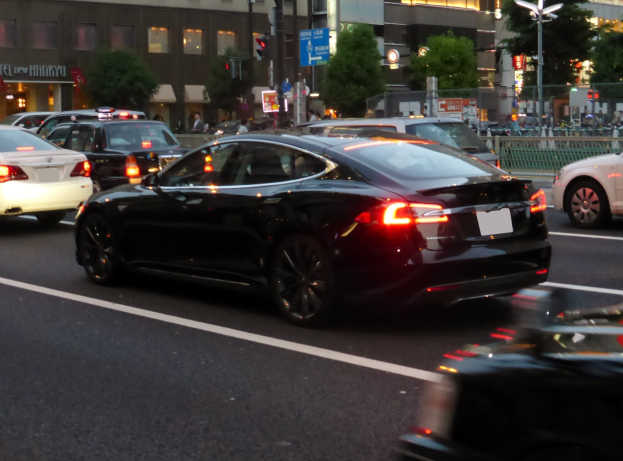Schwarzer Tesla Model S fährt auf einer Stadtstraße bei Nacht mit anderen Fahrzeugen, Gebäuden, Bäumen, Laternen, Verkehrszeichen, Geländern und Passanten im Hintergrund.