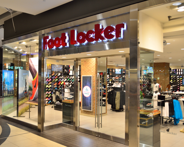 Ein Foot Locker Store in einem Einkaufszentrum mit Glaswänden und -türen, Kleider an Bügeln, Schaufensterpuppen in Kleidern, Texttafeln, Deckenleuchten und andere Gegenstände.