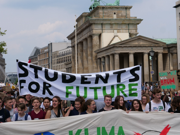 Eine Gruppe von Schülern marschiert in Berlin, eine bunt bemalte 'Students for Future'-Schlagzeile haltend, vor einem Hintergrund aus Gebäuden, Bäumen und Himmel.