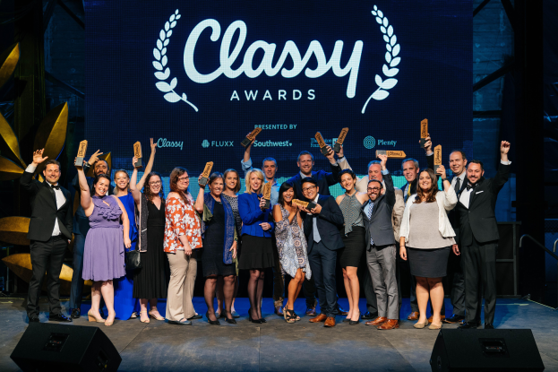 Eine Gruppe von Menschen auf einer Bühne, die lächeln und Preise halten, mit Lautsprechern unten und einem Bildschirm im Hintergrund, der "Classy Awards 2019" anzeigt.