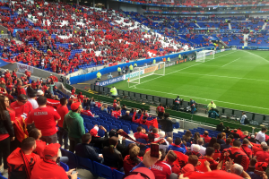 Eine große Menschenmenge in einem Stadion schaut ein Fußballspiel zu, mit Menschen auf Sitzen und Stehplätzen, Fahnen, einem Torpfosten und Deckenbeleuchtung unter einer elektrisierenden Atmosphäre.