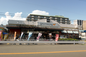 Eine belebte Stadtstraße mit Gebäuden, Straßenlaternen, Werbetafeln, Fahrzeugen, Absperrgittern, Büschen, Bäumen und einem bewölkten Himmel, mit einem Gourmet-Restaurant in der Mitte.