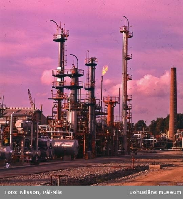 Ölraffinerie mit ausgedehnten Rohrleitungen, Metallkonstruktionen, Containern, Bäumen, einem Gebäude, einem Kran und einem bewölkten Himmel, mit Text am unteren Rand.