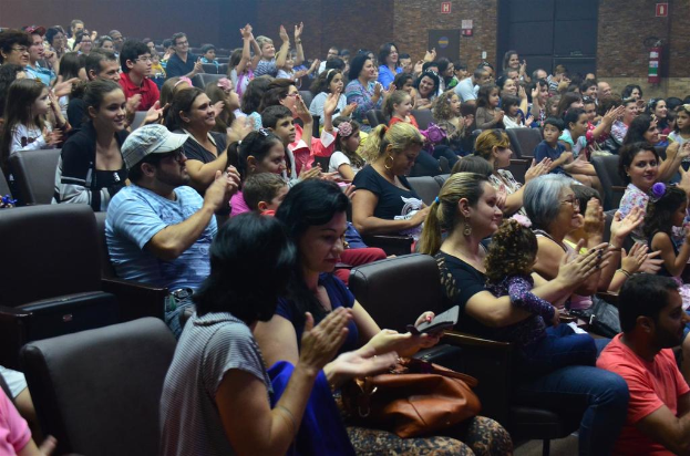 Eine Gruppe von Menschen, die auf Stühlen sitzen, lächeln, einige halten Gegenstände, tragen Brillen und einer trägt eine Mütze, während einige klatschen, mit einer Wand, Schildern und einem Feuerlöscher oben sichtbar.