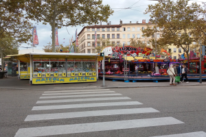 Eine belebte Stadtstraße mit Zebrastreifen und einer Karussellfahrt, mit Menschen, Buden, Bäumen, Gebäuden, Bannern, Laternenmasten und einer Ampel unter einem bewölkten Himmel.
