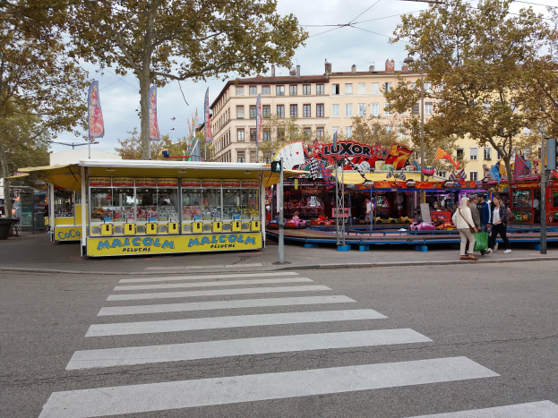 Eine belebte Stadtstraße mit Zebrastreifen und einer Karussellfahrt, mit Menschen, Buden, Bäumen, Gebäuden, Bannern, Laternenmasten und einer Ampel unter einem bewölkten Himmel.