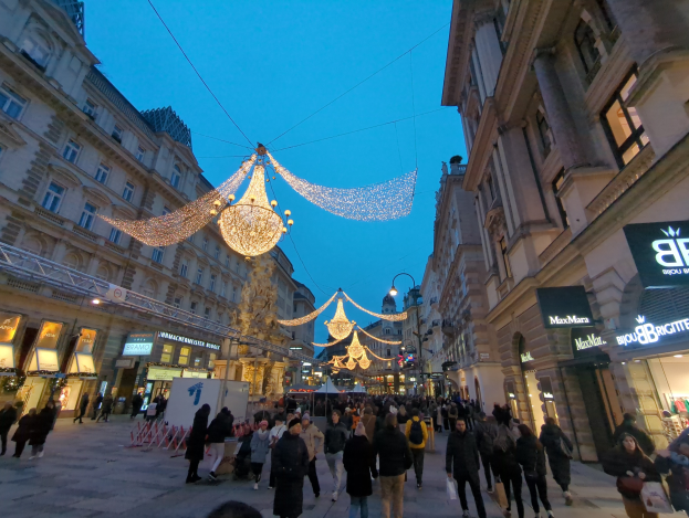 Eine belebte Stadtstraße voller Menschen, die unter festlichen Weihnachtslichtern spazieren, die von der Decke hängen, mit Gebäuden auf beiden Seiten und einem klaren blauen Himmel im Hintergrund.