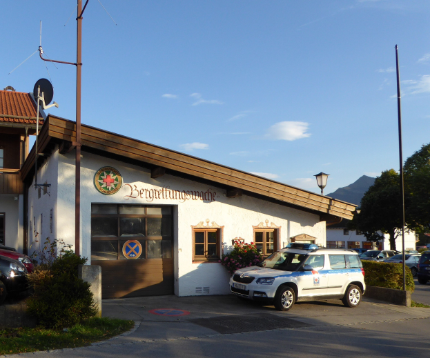 Polizeiauto vor einem Gebäude mit Fenstern und Türen geparkt, umgeben von Grün, Straßenlaternen und einem bewölkten Himmel.