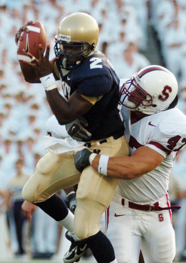 Zwei Fußballspieler in Helmen und Uniformen, einer fängt einen Ball in der Luft, während er von einem anderen getackelt wird, mit einem unscharfen Hintergrund.