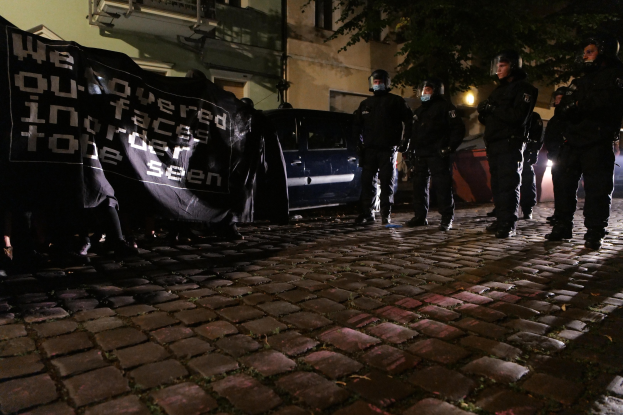 Eine Gruppe von Polizisten in Helmen steht auf einer Kopfsteinpflasterstraße bei Nacht, hält ein Banner und ist von einem Licht beleuchtet, mit Fahrzeugen, Gebäuden und Bäumen im Hintergrund.