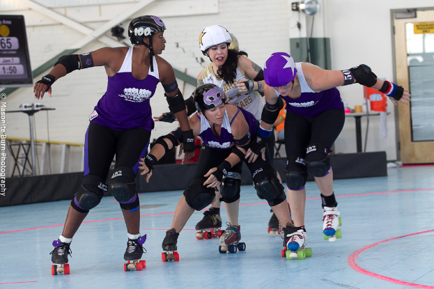 Vier Personen mit Helmen fahren in der Mitte, mit einer Wand, einer Tür, einem Stand, einem Mikrofon, Bannern, einem Tisch und anderen Gegenständen im Hintergrund.