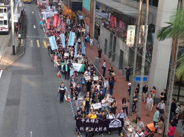 Eine große Gruppe von Menschen marschiert auf einer Straße in Hong Kong, hält Schilder und Plakate, mit Bäumen, gläsernen Gebäuden, Fahrzeugen und Schildern im Hintergrund.