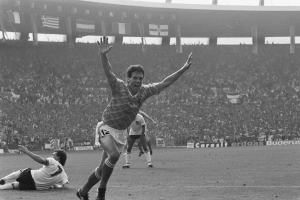 Ein Schwarz-Weiß-Foto eines Fußballspielers, der nach einem Tor feiert, umgeben von Menschen auf dem Boden, mit Brettern, Fahnen und Pfählen im Hintergrund unter einem klaren Himmel.