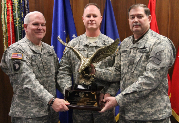 Drei Männer in Uniformen halten Auszeichnungen, mit Fahnen im Hintergrund und bunten Gegenständen mit Text oben links.