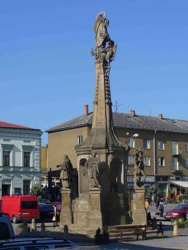 Ein Denkmal steht in einem Stadtplatz umgeben von Gebäuden, Straßenmöbeln, Fahrzeugen, Fußgängern, Bänken und Pflanzen unter einem bewölkten Himmel.