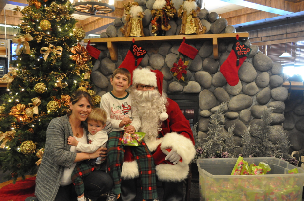 Eine Frau, Kinder und ein Mann in Santa-Klaus-Kostüm mit Brille und Handschuhen stehen neben einem geschmückten Weihnachtsbaum, einer Wand mit Felsen, Spielzeug, Dekorationen und einem Korb mit Gegenständen, mit Lichtern im Hintergrund.