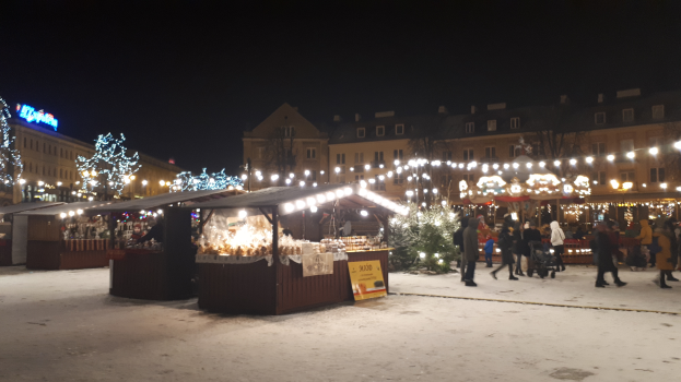 Ein nächtlicher Weihnachtsmarkt auf einer verschneiten Straße mit Menschen, Kinderwagen, beleuchteten Hütten, Pflanzen, Bäumen, Gebäuden und Schildern unter einem bewölkten Himmel.