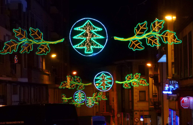 Eine städtische Straße, geschmückt mit festlichen Weihnachtslichtern, die Blätter und andere dekorative Elemente aufweisen, mit fahrenden Fahrzeugen und Gebäuden auf beiden Seiten.
