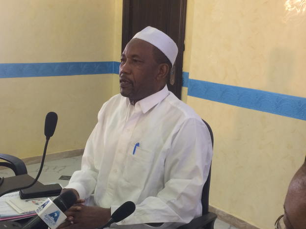 Ein Mann in weißem Anzug und Hut sitzt an einem Tisch mit Mikrofonen, spricht, während Papiere und Gegenstände auf dem Tisch zu sehen sind, mit einem anderen Mann mit Brille rechts von ihm in einem Raum mit einer Wand und einer Tür dahinter.