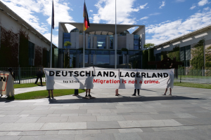 Gruppe von Menschen, die eine Fahne mit der Aufschrift "Deutschland, Lagerland Migration ist kein Verbrechen" vor einem Gebäude mit Fenstern halten, umgeben von Gras, einem Metallzaun, Pflanzen, Bäumen und Fahnenmasten.