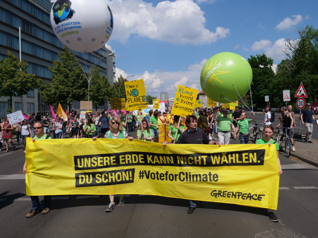 Eine Gruppe von Menschen hält ein gelbes "Wähle für das Klima"-Schild, während sie eine Straße mit Luftballons, Schildern, Fahrrädern, Bäumen, Laternenpfählen und Gebäuden im Hintergrund unter einem klaren blauen Himmel entlanggehen.