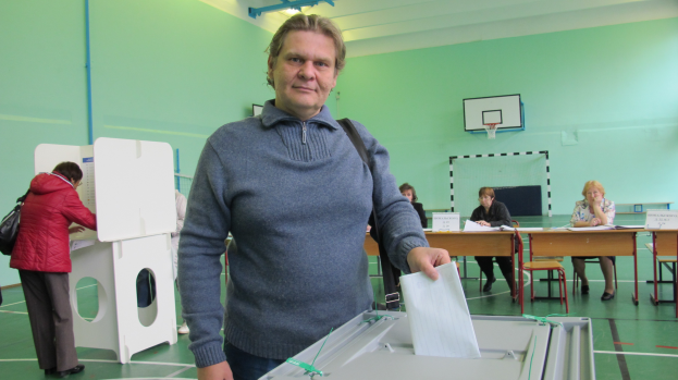 Ein Mann steht vor einer Wahlurne und hält ein Blatt in der Hand, umgeben von mehreren sitzenden und stehenden Menschen und einem Basketballkorb an der Wand im Hintergrund.