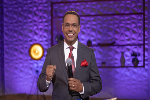 Stephen Colbert in Anzug, Krawatte und Brille auf einer Bühne stehend, ein Mikrofon haltend, mit beleuchtetem Ständer und verschiedenen Gegenständen dahinter.