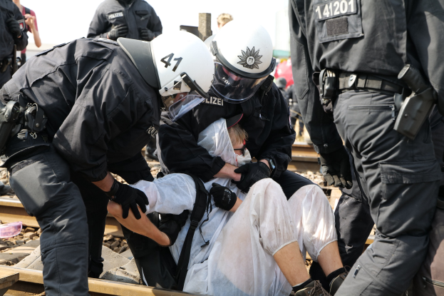 Eine Gruppe von Polizisten in Einsatzkleidung nimmt einen Mann in einem weißen Kleid auf einem Bahngleis fest, mit anderen Menschen im Hintergrund.