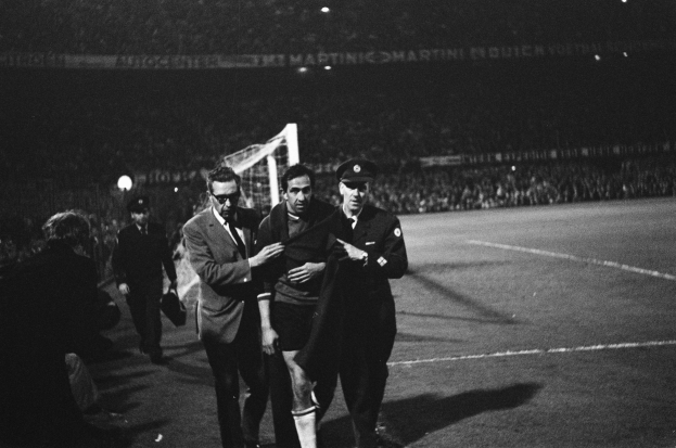 Eine Gruppe von Männern, die auf einem Fußballfeld feiern, mit einem Torpfosten und beleuchteten Tribünen im Hintergrund, dargestellt in Schwarz-Weiß.