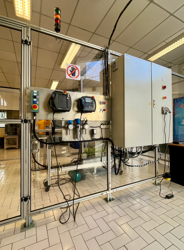 Eine große Maschine in einem Gebäude mit einem Boden, einer Wand, einer Decke mit Lampen und einer Tafel mit Text auf der rechten Seite, Teil eines Automatisierungssystems mit verschiedenen angeschlossenen Komponenten und Werkzeugen.