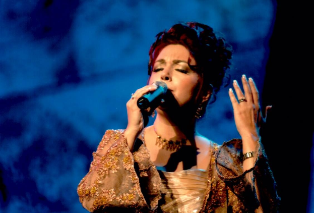 Frau in goldenem Kleid singt auf der Bühne der Eurovision Song Contest in ein Mikrofon mit blauem und schwarzem Hintergrund.