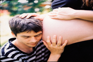 Ein Mann und eine Frau stehen nebeneinander, der Mann berührt zart den Bauch der Frau, mit einem unscharfen Hintergrund, der das Paar und ihr ungeborenes Kind betont.