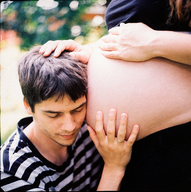 Ein Mann und eine Frau stehen nebeneinander, der Mann berührt zart den Bauch der Frau, mit einem unscharfen Hintergrund, der das Paar und ihr ungeborenes Kind betont.