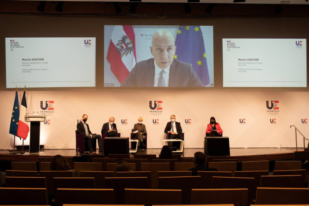 Eine Gruppe von Menschen, die auf einer Bühne in einem Halbkreis um ein zentrales Podium mit Mikrofon sitzen, mit Flaggen auf beiden Seiten und einem Bildschirm mit Text und Bildern im Hintergrund, während einer Pressekonferenz der Europäischen Union.