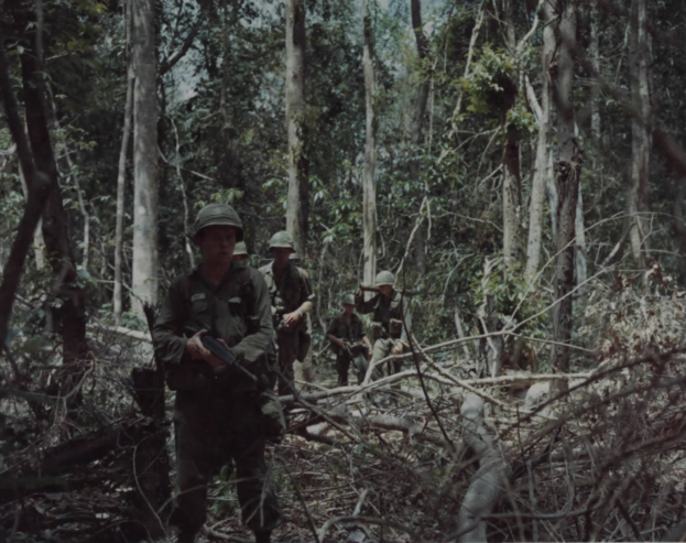Eine Gruppe von Soldaten in Helmen und mit Gewehren marschiert durch einen dichten Wald mit hohen Bäumen und einem sichtbaren Himmel, mit Text am unteren Bildrand.