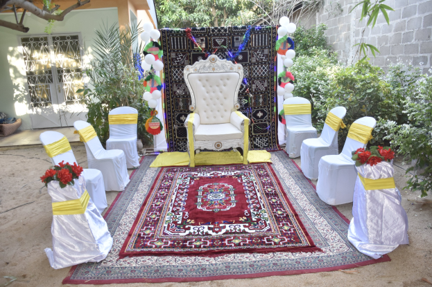 Hochzeitszeremonie im Garten mit weißen Stühlen, einem roten Teppich, Blumenbouquets, Dekorationsgegenständen, Luftballons, Pflanzen, Bäumen, einem Gebäude mit Fenstern und einer Tür sowie einer Wand auf der rechten Seite.