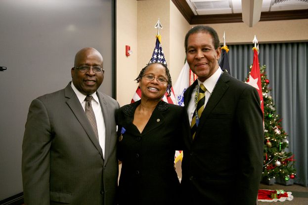 Eine Gruppe strahlender Menschen vor einem geschmückten Weihnachtsbaum mit einem Vorhang im Hintergrund, Flaggen und verschiedenen Gegenständen, mit einem Tuch und mehr Gegenständen auf der rechten Seite.
