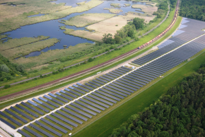 Luftaufnahme einer Solar-Farm mit Panelen, umgeben von Bäumen, Gras, Wasser und einer Bahn auf einer nahen Bahnstrecke.