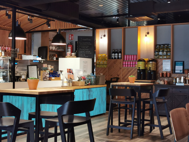 Inneneinrichtung eines Restaurants mit Holztischen, -stühlen, einem Blumentopf auf einem Tisch, einer Schiefertafel, einem Kühlschrank, einem Feuerlöscher, Regalen mit Flaschen, Schränken mit Gegenständen, einem Rahmenfoto an der Wand und Deckenleuchten.