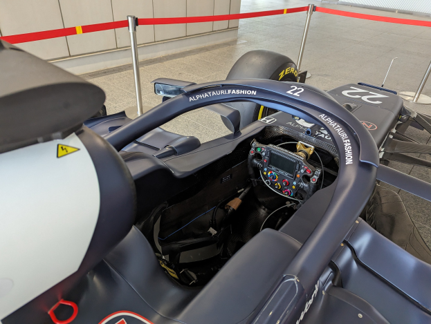 Cockpit eines Formel-E-Autos in einer Ausstellungshalle mit einem Lenkrad im Vordergrund, einer Wand im Hintergrund und um das Auto herum abgesteckte Pfosten.