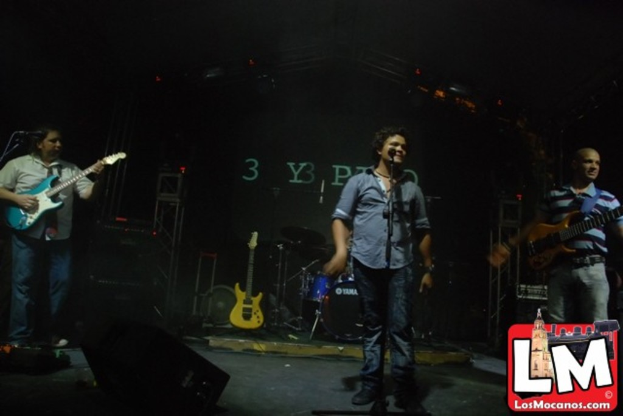 Drei Personen auf der Bühne, eine spielt Gitarre und eine andere singt in ein Mikrofon, mit einem Snare-Drum-Arrangement und Bühnenbeleuchtung im Hintergrund.