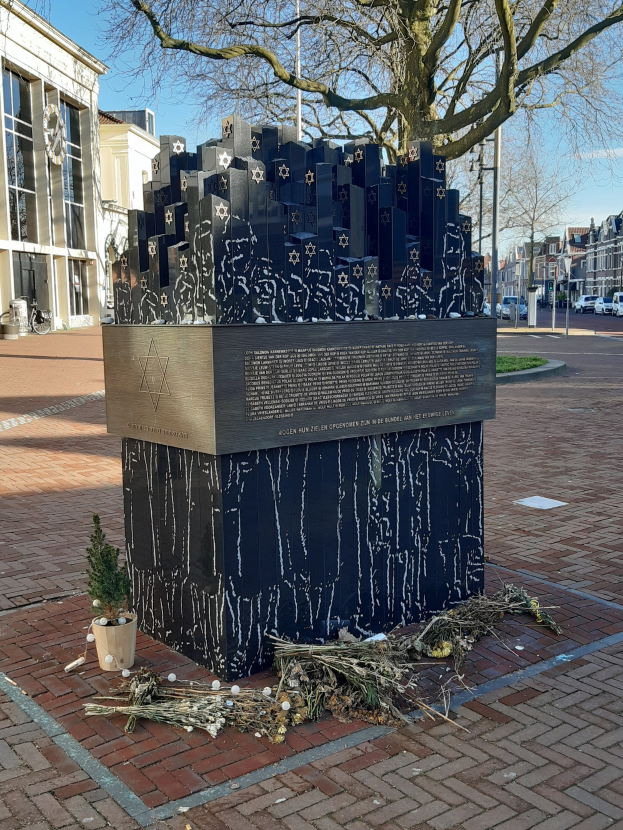 Denkmal in einem Stadtplatz für Holocaust-Opfer mit einem Baum davor, einem Topf mit einer Pflanze, verstreuten trockenen Blättern und Gebäuden, Fahrzeugen, Pfählen, Bäumen und Himmel im Hintergrund.
