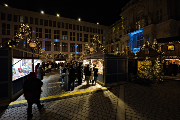 Ein belebter Weihnachtsmarkt bei Nacht mit Menschen um geschmückte Stände, festlicher Beleuchtung und Gebäuden im Hintergrund unter einem dunklen Himmel.