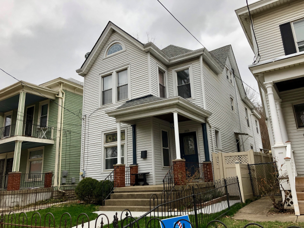 Reihe von Häusern mit Fenstern, Türen, Säulen, Geländern, Stufen und Dächern, umgeben von einem Metallzaun, Gras, Pflanzen, Bäumen und einem bewölkten Himmel, mit einem Mietschild am Boden, das die Adresse und die Listen-ID auflistet.