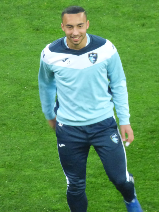 Ein Mann in einem blauen Hemd und schwarzen Hosen lächelt, während er auf einem Fußballfeld läuft und Schuhe trägt.