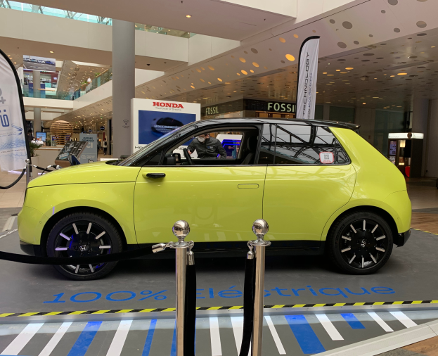 Ein gelbes wasserstoffbetriebenes Elektroauto steht vor einem Gebäude, umgeben von Absperrpfosten mit Seilen, Schildern und Texttafeln, mit einer Person im Auto und einer Gruppe von Menschen in der Nähe, unter Deckenleuchten und Ventilatoren.