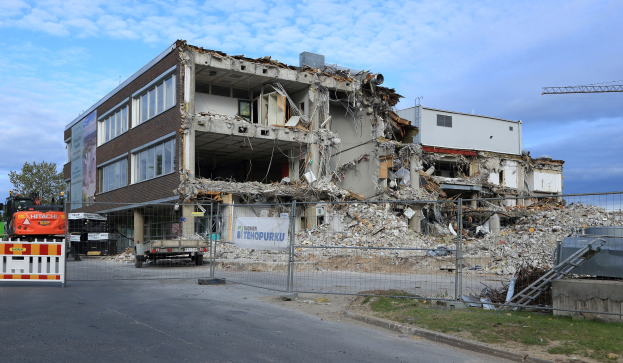 Ein großes Gebäude wird in der Mitte einer Straße abgerissen, umgeben von einem Metallzaun, Fahrzeugen, Gras, einem Schild, einer Fahne, Pfählen, einem Kran, Bäumen und einem bewölkten Himmel.
