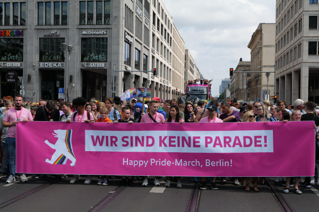 Eine Gruppe von Menschen marschiert auf einer Straße in Berlin, Deutschland, mit einer pinken 'Happy Pride March'-Tafel, mit Gebäuden, Laternenmasten und Verkehrszeichen an der Straße.
