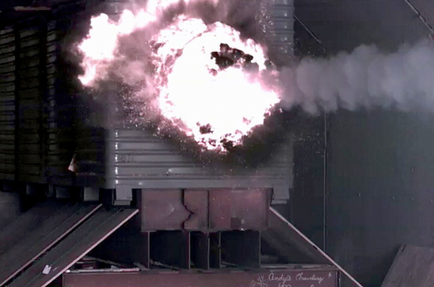Große Explosion mit Rauch, der aus einem Gebäude aufsteigt, eine Wand im Hintergrund und verstreute Metallobjekte und eine Texttafel unten.