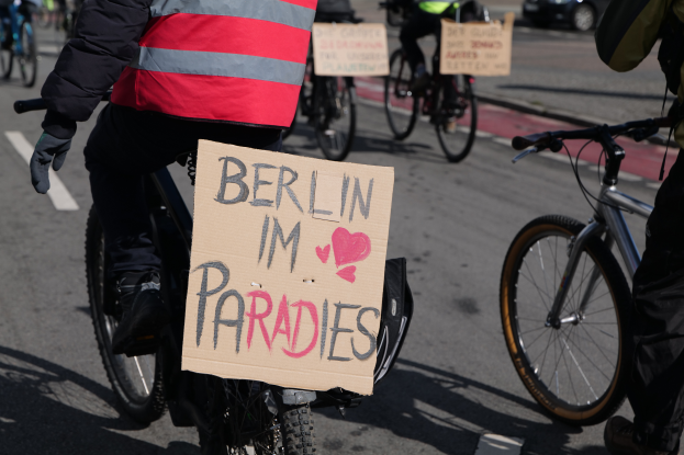 Eine Gruppe von Menschen, die auf Fahrr√°dern eine Stra√e entlangfahren, mit einem Schild "Berlin I'm Paradies" im Vordergrund, einem Auto im Hintergrund und dem Bild ist leicht unscharf.